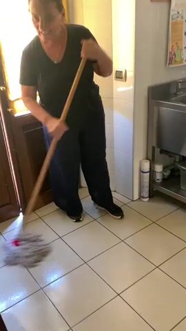 A woman is mopping a tiled floor in what appears to be a kitchen or food preparation area. She is wearing a dark t-shirt and dark, wide-legged pants, and black sneakers. She is holding a mop with a long wooden handle and a round mop head, which she is using to clean the floor. The floor is light-colored tile, and appears wet. In the background, there is a stainless steel sink and counter area, as well as what looks like kitchen equipment. There is also a door on the left side of the frame with a yellow curtain. The lighting suggests it is daytime, and the environment appears to be indoors.