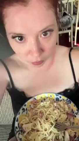 A woman with red hair eats a bowl of spaghetti. She wears a black tank top. She uses a fork to lift spaghetti into her mouth. The bowl has a blue, yellow and white pattern. The video is taken from a close-up angle. A dog is visible in the background.