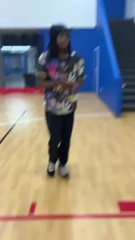 The video shows a person throwing a basketball into a hoop. The person is male, has long dark hair, is wearing a black beanie, a t-shirt with a colorful pattern, black sweatpants, and black and gray shoes. The person is standing on a basketball court in an indoor gym. The floor is shiny wood, and the walls are white and blue. There are stairs in the background and windows near the ceiling. The person throws the basketball and it goes through the hoop and bounces onto the floor.