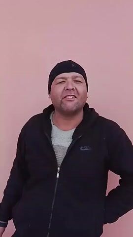 A medium shot shows a man standing in front of a plain pink wall. He wears a black beanie, a black zip-up hoodie with a Nike logo, and a gray shirt underneath. He has a slight smile and is looking directly at the camera. In the beginning, he says a word, then starts to laugh. He then says another phrase. He has his hand on his hip and brings out a cigarette to smoke.