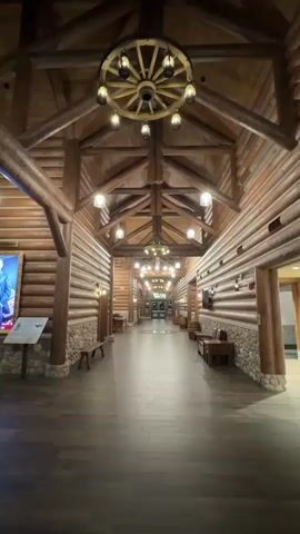 The video shows a long, wide hallway in a rustic, log-cabin-style building. The walls and ceiling are constructed from large, round logs, and the ceiling features exposed wooden beams and rafters. A large wagon wheel-shaped chandelier with multiple lights hangs from the ceiling near the beginning of the hallway. Along the sides of the hallway, there are stone bases and more log construction. Several smaller, rustic-style light fixtures are mounted on the walls. On the left wall, there is a framed portrait of a man in a suit, with a small information plaque below it. There are also a few wooden benches along the walls. The floor is made of dark wood planks. The hallway extends into the distance, where more lights and chandeliers are visible, suggesting it leads to other areas of the building. The overall impression is one of a grand, log-themed interior, possibly a lodge, hotel, or public building. No people are visible in the hallway during this short clip.