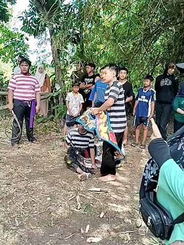 The scene depicts a group of people gathered in a wooded area, seemingly observing a cultural performance.  A young man wearing a striped shirt is the main focus, as he manipulates a colorful, fringed object while kneeling on the ground. Several other people, including children and adults, are standing around watching.  The environment appears to be a rural area with trees and dirt ground. A few structures, possibly small buildings or fences, are visible in the background. The people's clothing ranges from casual attire to traditional garments.