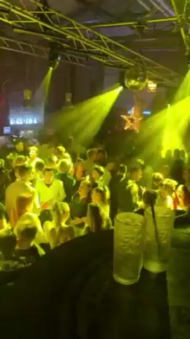 The video shows a crowded nightclub illuminated by yellow stage lights and a disco ball. Many people are standing close together and possibly dancing. Two tall glasses with straws are in the foreground, possibly on a bar. The building has exposed metal beams and industrial-style lighting fixtures. The general atmosphere is lively and energetic.