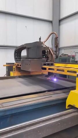 A close-up, medium shot captures a plasma cutter in action within a factory setting. The machine, predominantly yellow and dark gray, features a gantry system with a plasma torch that is actively cutting through a large, flat sheet of dark metal. Sparks and molten material fly from the point of contact as the torch moves from right to left across the metal. The torch is connected by a flexible conduit to a boxy component above, which is part of the gantry. A visible chain-like mechanism runs along the gantry, likely for controlled movement. The background reveals the industrial environment with corrugated metal walls and a large vertical pillar. The lighting is functional, illuminating the workspace. No people are visible.