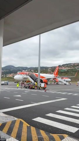 The video shows three planes parked on a runway under a cloudy sky. The first plane, an EasyJet aircraft, is orange and white, and the second, a Wizz Air plane, is white and pink. The third plane is a Ryanair plane. Airport personnel, wearing reflective vests, are working around the EasyJet and Wizz Air planes. The airport buildings are partially visible in the foreground, and a hill dotted with houses is visible in the background.