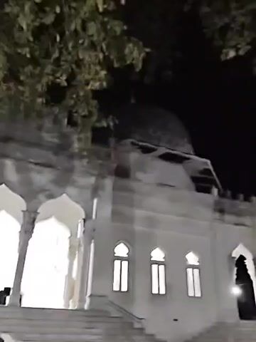 The video depicts a white mosque at night. The camera angle is from below, looking up at the mosque's facade. Three arched entrances are visible, illuminated from within, with white pillars on either side of each arch. To the side, three arched windows are seen. The mosque has a dome at the top. A tree with leaves obscures some of the view above the mosque. A motorcycle is parked in front of the steps leading up to the mosque. The background is dark, suggesting it's nighttime.