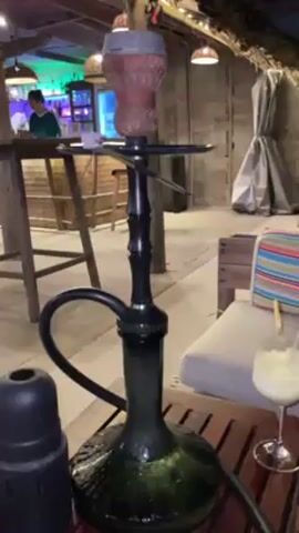 Here's a description of the video:
The video shows a hookah set up on a wooden table.  The hookah is black, with a green-tinted glass base. A black hose extends from the hookah. A black charcoal holder sits to the left of the hookah. Next to the hookah is a glass containing a white, frozen-looking drink with a lemon wedge and two straws. A striped pillow sits on a cushioned bench behind the glass. The background shows a bar area with colorful lights and a person standing behind the bar. The overall setting appears to be an outdoor lounge area, possibly at a resort or restaurant. The video appears to be stationary, focusing on the hookah.