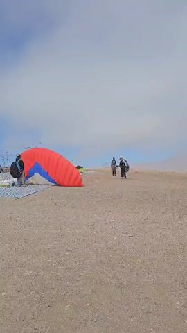 The scene is set on a sandy, possibly desert-like terrain under a sky dominated by light blue with a veil of white clouds. There are four individuals present in the video. One is near a large, partially inflated paragliding parachute that is predominantly red with a patch of blue; this person appears to be readying the equipment. Three individuals walk into the frame from the right. They are dressed in outdoor attire, including helmets and backpacks. One wears sunglasses. The general activity suggests preparation for a paragliding event, with people and equipment being readied on the ground.