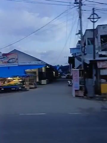 The video shows a street scene at dusk or dawn. There are several market stalls and shops lining the road, many with blue tarps overhead. Some stalls appear to be selling food, with cooking equipment visible. A few people are present, some standing near the stalls, and one appears to be on a motorcycle. A yellow truck is parked at one of the stalls. The sky is cloudy.