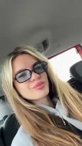 A woman with long blonde hair is sitting in the passenger seat of a car. She is wearing glasses and looking towards the camera. The interior of the car is visible, and the background is blurred. The lighting is natural, and the overall scene appears casual.