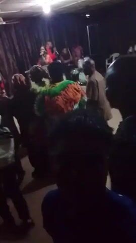 The video depicts a dimly lit indoor space with a gathering of people. The scene appears to be a social event or celebration, as indicated by the music and the vibrant clothing worn by some individuals. People are dancing and moving around. The room contains chairs, tables, and a stage in the background. The video is recorded from the perspective of someone moving through the crowd.