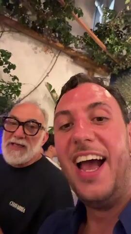 The video shows a selfie-style recording of two men in what appears to be an indoor dining setting. In the foreground, a man with short, dark curly hair and a bright red complexion is speaking directly to the camera, his mouth open. He is wearing a dark blue collared shirt. Behind him and to the left, an older man with gray hair and a beard, wearing glasses and a black t-shirt with white text, is visible. He is smiling and looking towards the camera. Other people and foliage are blurred in the background, suggesting a restaurant or social gathering. The lighting is warm.