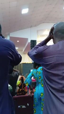 The video shows several people gathered in what appears to be a church or large hall. The people are dressed in colorful, patterned clothing, and some are raising their hands in the air. The camera focuses on the backs of the people in the foreground, while others can be seen in the background. Overhead lighting illuminates the scene, and there are large speakers hanging from the ceiling. The people seem to be engaged in a worship or prayer service.