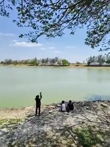 The video shows a group of people by a body of water. In the foreground, a person in a dark hooded jacket stands on the bank of a light green body of water, facing away from the camera and holding what appears to be a long object, possibly a fishing rod. To the right of this person, two other individuals are seated on the ground. The terrain is rocky and somewhat dry, with patches of grass and scattered debris. Trees with sparse foliage hang over the scene from the top of the frame, creating dappled shade. In the background, the water stretches to a distant shoreline lined with trees and some structures, under a bright blue sky with scattered white clouds. The overall scene suggests an outdoor recreational setting.