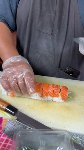 A person wearing a dark apron and clear plastic gloves is preparing sushi. They are standing in front of a white cutting board where a cut sushi roll, topped with slices of salmon, is placed. A knife with a wooden handle rests to the left of the cutting board. To the right of the cutting board, there are several clear plastic containers, some filled with sliced salmon and others with julienned cucumber. The person is holding another clear plastic container, appearing to be assembling packaging. The scene appears to be outdoors, possibly in a market or food stall, given the lighting and the presence of other containers and what might be a chair in the background. The overall impression is of food preparation in a casual setting, likely a street food vendor or a small restaurant in Ko Samui, Thailand. There is no indication of the time of day or weather, and no visible emotions are discernible. Text is visible on the apron in green letters, but it is partially obscured and difficult to read clearly.