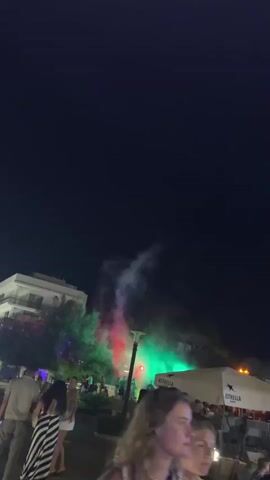The video shows a nighttime scene with several people walking around an outdoor area. There are buildings in the background and trees with colored lights shining on them. In the center of the frame, there is a fountain or mist effect with red and green lights. Some people are sitting at tables under umbrellas. The overall atmosphere appears to be festive.
