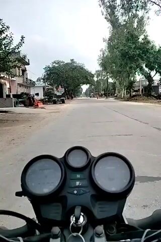 The video is a first-person perspective from a motorcycle, showing the rider's view of a road with buildings and trees on either side. The motorcycle's handlebars and dashboard, including the speedometer and tachometer, are visible in the foreground. The road appears to be paved and in a rural or suburban setting, with a few scattered vehicles and people in the distance. The sky is overcast, suggesting a cloudy day. The rider is moving forward at a steady pace.