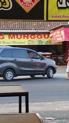 A grey car is driving down a street. To the right of the car, a person wearing a white t-shirt and shorts is standing and holding a white object. Behind the person is a pink banner that says BEBEK GORENG SERUNDENG "MAS ANTON". There are also several motorcycles parked on the side of the road.