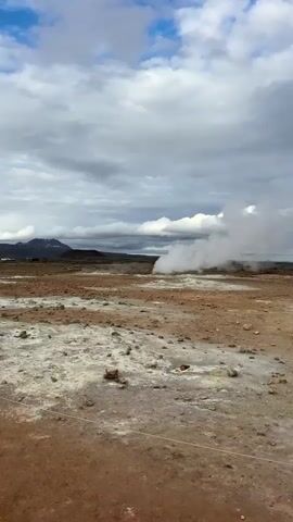 The video captures a wide shot of a barren, geothermal landscape in Reykjahlíð, Iceland, under a partly cloudy sky with patches of blue. The ground is a mix of reddish-brown earth and lighter, yellowish-white mineral deposits, with scattered rocks throughout. In the mid-ground, a powerful plume of white steam and gas rises significantly into the air from a geothermal vent, indicating active volcanic or geothermal activity. Low-lying hills or mountains are visible in the distant background. No people or text are visible in the frame. The scene conveys a sense of raw, natural power and a stark, unique environment.
