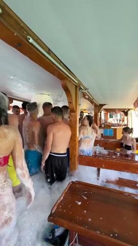 The video captures a lively foam party taking place inside a boat, likely a party cruise or tour boat. Numerous young adults, both male and female, are seen dancing and standing in a thick layer of white foam that covers the boat's floor.

Most of the men are shirtless, wearing swim shorts in various colors like yellow, blue, and black. The women are dressed in swimwear, predominantly bikinis, with one prominent figure in a red bikini top and pink bottoms. People are fully immersed in the foam, which reaches up to their waists in some areas.

The boat's interior features wooden beams, pillars, and structural elements, creating a rustic yet festive atmosphere. Wooden tables and benches are visible on the right side of the frame, some occupied by people sitting, others covered in foam. In the background, a bar area is discernable with bottles and glasses, indicating where drinks are served. The ceiling is white, and the overall lighting suggests it's daytime, possibly on a sunny day given the nature of the event.

The participants appear to be in high spirits, enjoying the music and the unique experience of the foam party. Their body language suggests dancing, interaction, and general revelry. No discernible text is visible on clothing, signs, or objects within the frame. The scene conveys a fun and energetic vacation or leisure activity.
