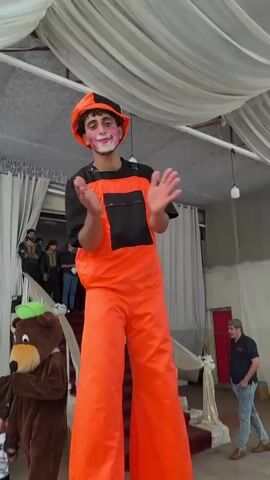 The video shows a lively event taking place indoors, likely a celebration or performance, under a white draped canopy. The main subject is a performer on stilts, dressed in bright orange overalls over a black t-shirt and a red cap. The performer has clown-like makeup with pink and purple accents on their cheeks and around their mouth, and is gesturing with their arms, appearing to engage with the audience.

To the left, a person in a brown bear costume with a green hat stands next to a young child wearing a striped shirt. In the background, there are people seated on white chairs, some wearing headscarves. A red carpeted staircase is visible behind the bear character, with several people standing at the top. Another man in a light blue shirt and jeans is seen to the right, facing away from the camera.

The overall atmosphere seems festive, with the bright colors of the performer's costume and the draped ceiling creating a celebratory mood. The lighting suggests it is daytime, but the indoor setting makes it difficult to determine the exact time of day or weather. The presence of a seated audience and performers in costumes points to an organized event, possibly a party or a community gathering. There is no visible text in the video.