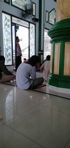 The video shows an interior view of what appears to be a mosque or a similar place of worship. Several individuals are engaged in prayer, kneeling on prayer mats. The floor is tiled, and there are large decorative pillars with green and cream colored accents. Windows with decorative stained glass patterns are visible, letting in natural light. In the foreground, one person is seen from behind, wearing a checkered shirt and dark pants, kneeling on a prayer mat. Further back, other individuals in white garments are also in prayer positions. The overall atmosphere suggests a religious gathering or service.