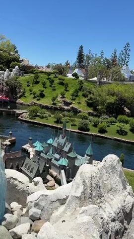 This video captures a bright, sunny day with a clear blue sky, likely in a theme park. The footage shows a miniature village with tiny houses and a castle with green roofs. A river flows through the village, and a small boat with several people is visible on the water, entering a tunnel. The surrounding landscape is lush and green, with manicured trees and bushes, and rocky, mountainous features. The camera pans across the scene, offering different perspectives of the miniature world. The video briefly cuts to a view from inside a vehicle, looking out at a stone bridge and train tracks.