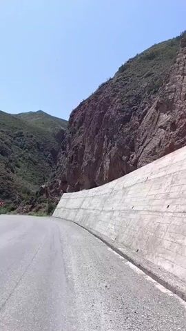 A road curves through a mountainous landscape under a clear blue sky. On the right, a steep, rocky cliff face rises dramatically, with a reinforced concrete wall supporting the road's edge. To the left, a gentler, vegetated slope climbs upwards, dotted with sparse greenery and rocks. In the distance, more rugged mountains stretch into the hazy horizon. Several red and white directional signs are visible on the left side of the road, indicating upcoming turns. The asphalt road appears to be in good condition, with faint tire marks visible. The overall impression is one of a remote and scenic route, likely in a dry, mountainous region.