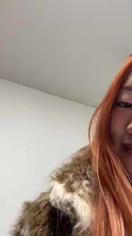 An Asian woman with orange-red hair, wearing a fuzzy coat, extends her hand towards the camera. She makes scratching sounds with the extremely long, pointed white nails on that hand. The background appears to be a white wall.