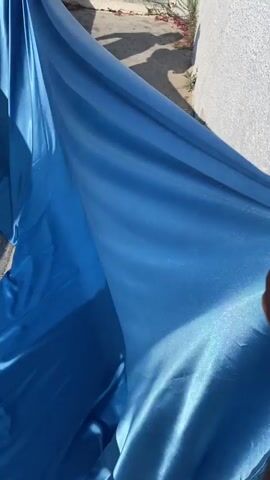 The video opens with a close-up of a shimmering blue fabric, likely a dress, draped over a surface. The camera then pans out to reveal a couple posing for a photo against a backdrop of white buildings and the blue sea, characteristic of a Mediterranean island like Santorini. The man, wearing a white shirt and shorts, leans against a white railing. The woman is wearing a flowing blue gown with ruffled sleeves and a long train that spreads across the ground. The couple is positioned on a balcony or terrace overlooking the ocean. The latter part of the video shows the scenic view of the ocean and the cliffside village.