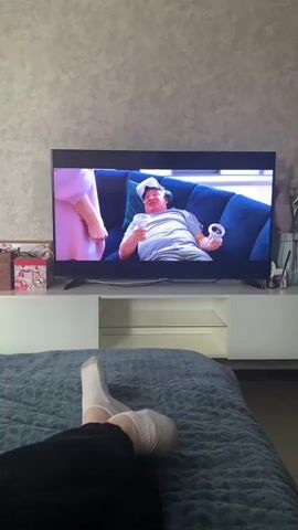 A person is lying on a bed, wearing socks and watching a TV. The TV is showing a scene with a man lying on a blue couch wearing a VR headset and holding controllers, and a woman looking at him with a disapproving expression. The scene then cuts to the woman standing in a large, bright living room. The TV is on a white media console with a glass shelf. A box and vase are on the side of the console.