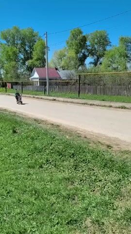 The video shows a young person riding a small, orange motorbike down a dirt road in a rural setting. The rider is wearing a blue and green hooded jacket and black pants. The road is bordered by a grassy area and a fence, with trees and houses visible in the background. The sky is clear and blue. The rider passes by the camera.