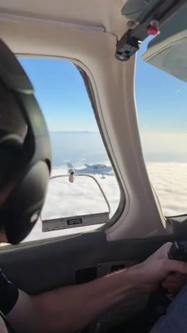 A white Cirrus aircraft is flying above clouds. The camera appears to be from another aircraft or a drone, looking at the Cirrus from an angle. The pilot can be seen inside the cockpit. The sun is shining, creating a bright, slightly hazy atmosphere. The Cirrus aircraft has its landing gear extended. The propeller is visible and appears to be spinning.
