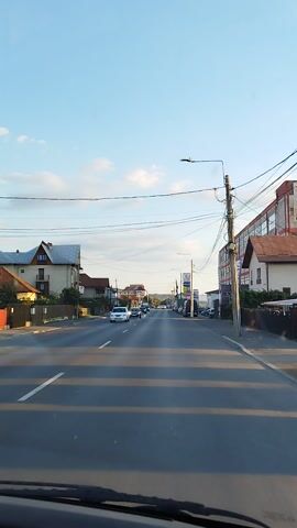 This media file shows a view from a vehicle traveling on a street in Curtea de Argeș, Romania. The scene depicts a moderately busy road with several cars driving in the same direction as the camera. The time of day appears to be late afternoon or early evening, given the angle of the sun and the soft lighting. The sky is clear with a few scattered clouds.

Along the street, there are buildings on both sides. On the left, a large, multi-story building with a red and white facade is prominent, possibly a commercial or industrial structure. Further down the street on the left, there are other buildings and signs, including one that appears to be for "AVISTA". Utility poles with a complex network of wires stretch across the scene. On the right side of the road, there are residential houses with pitched roofs.

The activity captured is vehicular traffic, with cars and a truck visible. There are no people clearly discernible in the foreground or walking on the street in a way that draws attention. The overall atmosphere seems calm and ordinary, typical of a suburban or semi-urban environment. The image quality is decent, though slightly tilted. The focus is on the road and the surrounding environment, giving a sense of journey or travel through this Romanian town.