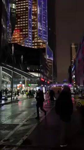 The video depicts a city street at night. The street is wet, and the reflections from the streetlights and building lights are visible. Tall buildings loom over the street, their lights glowing in the darkness. One building is particularly prominent, featuring a distinctive design and lit with blue accents. People are walking along the street, some individually and some in groups. The overall atmosphere is urban, modern, and somewhat busy despite the late hour.