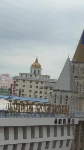 A video shows a cityscape with several buildings. In the foreground, there is a building with a white facade and columns. The middle ground features a building with a tall, conical tower and several smaller pointed roofs. In the background, a large, light-colored building with a golden dome is visible, along with other residential buildings and industrial chimneys. The sky is overcast.