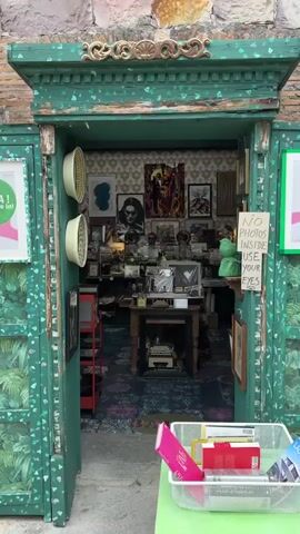 The camera slowly zooms in on the interior of a store. The doorway is painted green and decorated with gold accents and leaf patterns. On the left, there is a sign with a floral design. On the right, there is a sign that reads "NO PHOTOS INSIDE USE YOUR EYES." Inside the store, there is a collection of objects including artwork, photographs, vintage items, and books. A vintage typewriter sits on a table in the foreground. The floor is decorated with a colorful, intricate pattern. The walls are covered in art and the shelves are filled with various items. Track lighting can be seen on the ceiling.