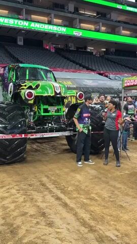 A medium shot captures a monster truck show inside an arena. The monster truck, "Grave Digger," is positioned on the dirt floor, its prominent green and black exterior with flame decals visible. Two individuals are standing in front of the truck, engaged in a conversation. One, wearing a green and black uniform with "Grave Digger" branding, stands closer to the truck and appears to be speaking. The other, a woman in a red shirt and black leggings, holds a microphone and is facing the man, seemingly conducting an interview. The background reveals stadium seating, mostly empty, with a banner displaying "WESTON ANDERSON" and "GRAVE DIGGER." Other spectators can be seen further back, and a large tent-like structure is behind the truck. The lighting suggests an indoor event.