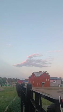 A vertical video shows a rural landscape under a pale blue sky with wispy pink clouds. In the foreground, a dark wooden fence runs horizontally across the lower part of the frame. Beyond the fence, a bright red, two-story building with a gray roof is centrally located. To the left of the building, there are glimpses of other structures and trees, suggesting a village or small town. The ground in front of the fence is covered in green grass. The lighting suggests it is either early morning or late evening.