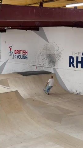 A man on a scooter performs a backflip on a halfpipe. He's wearing a white t-shirt and blue jeans. The halfpipe is made of wood and is located indoors. Behind the halfpipe is a white wall with the "British Cycling" logo. The man lands the backflip successfully and rides away.