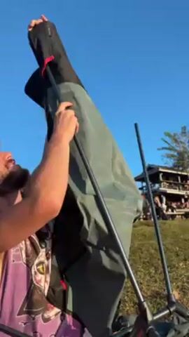 A man is seen setting up a portable camping chair outdoors, possibly in a park or at an event. The sky is a clear, bright blue. The man is wearing a purple tank top with a graphic on it. He has curly brown hair and a beard. He is holding the chair's frame in one hand and the fabric of the chair in the other. The chair is a black and gray color. The background shows a wooden building with people around, likely attendees or onlookers. The grass is green and dry. There are a few trees and what looks like a palm tree visible in the background as well.
