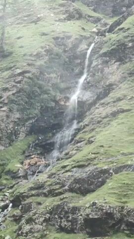 The video shows a narrow waterfall cascading down a rocky, moss-covered mountainside. The water flows in a white, frothy stream over dark, wet rocks. The surrounding slopes are covered in lush green grass and sparse vegetation. The scene appears to be in a natural, outdoor environment, likely a mountainous region. There are no people or man-made objects visible in the footage. The weather seems damp or rainy, as evidenced by the mist and water droplets in the air.