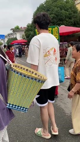 A street scene in what appears to be a market. In the foreground, a young man wearing a white t-shirt, black shorts, and white slides carries a large, woven basket on his left shoulder. A person in a purple top and plaid skirt is mostly visible on the left, also with a bag strap over their shoulder. An older woman in a patterned brown top and beige pants walks beside the young man, looking towards him. In the background, there are market stalls with red umbrellas, and other people can be seen walking. A sign with Chinese characters is visible behind the young man. The ground is paved and appears wet in some areas.