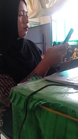 A person wearing a black hijab and a striped shirt is sitting at a table covered with a green cloth. They are holding a smartphone in their hands and looking at it. A laptop is open on the table in front of them. The background shows a window with green curtains and some objects on shelves. The lighting suggests an indoor environment, possibly a classroom or office.