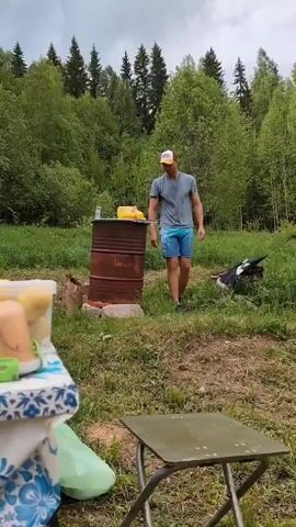 A man is standing near a rusty metal barrel in a grassy area with trees in the background. He is wearing a grey t-shirt, blue patterned shorts, a baseball cap, and black shoes. The barrel has a yellow object and a small bottle on top of it. In the foreground, there is a small metal table, a container of food wrapped in plastic, and what appears to be a green bag. The man makes a "peace" sign with his fingers while looking towards the camera. The day appears overcast.
