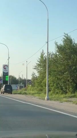The video shows a view from a vehicle traveling on a road. On the left side of the road, a tall sign with the logo "IRBIS" is visible, displaying fuel prices. The prices for different octane fuels (92, 95, 98, and DT) are shown as 00.00, indicating they are not being displayed or are set to zero. A silver SUV is parked near the gas station. Further down the road, a green gas station canopy and buildings are visible. The environment appears to be a semi-rural or suburban area with trees lining the right side of the road and a clear blue sky overhead. There are power lines crossing the scene. The camera is in motion, suggesting the video was recorded from a moving vehicle.