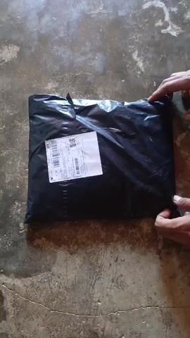 A pair of hands are shown reaching for a black plastic-wrapped package lying on a concrete surface. The package has a white shipping label affixed to it. The hands adjust the package.
