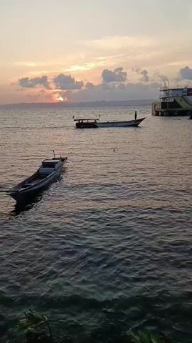 The video shows a body of water with several boats. In the foreground, a small, dark boat is visible on the left, partially submerged. In the middle ground, a longer, lighter-colored boat with an awning is moving from left to right. A person stands near the stern of this boat. In the background, a large white ship with yellow and red accents is docked or anchored. The sun is setting or rising, casting a warm glow on the sky, which is filled with clouds. The water is rippling. There is a shoreline visible on the far left.