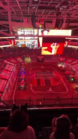 The video shows a high-angle view of a monster truck rally event taking place inside an arena. The arena is dimly lit, with predominantly red lighting casting a vibrant glow over the scene. In the foreground, the backs of two spectators are visible, one with what appears to be animal ears attached to their head. The main focus of the video is the dirt arena floor, which is marked with white lines indicating a track. Lined up on one side of the arena are several colorful monster trucks, each with unique designs and illuminated headlights. The trucks are positioned in a row, facing towards the center of the arena. In the background, a large screen displays an image of a person in a cowboy hat. Other screens and advertising banners are visible around the arena, along with glimpses of stadium seating filled with an audience. The overall atmosphere appears to be energetic and anticipatory, likely before the start of the event.
