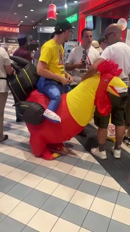 The video shows a person wearing an inflatable rooster costume in a public indoor space, possibly a retail store or a service area. The person is seated on the rooster's back, with their legs extending out from under the costume. They are wearing a yellow t-shirt, blue pants, and white sneakers. The rooster costume is predominantly red, with yellow and black accents. The person is holding onto the costume, and the rooster's head is to the right of the frame.

Several other people are visible in the background. To the left, a woman with blonde hair is partially visible. To the right, a man wearing a white t-shirt and shorts is standing and looking towards the person in the costume. Another person is partially visible behind him. The floor is tiled with a checkered pattern of white and light gray squares. In the background, there are shelves with products and signs. One sign reads "TOILETS." The lighting appears to be artificial. The overall atmosphere suggests a casual or festive setting.